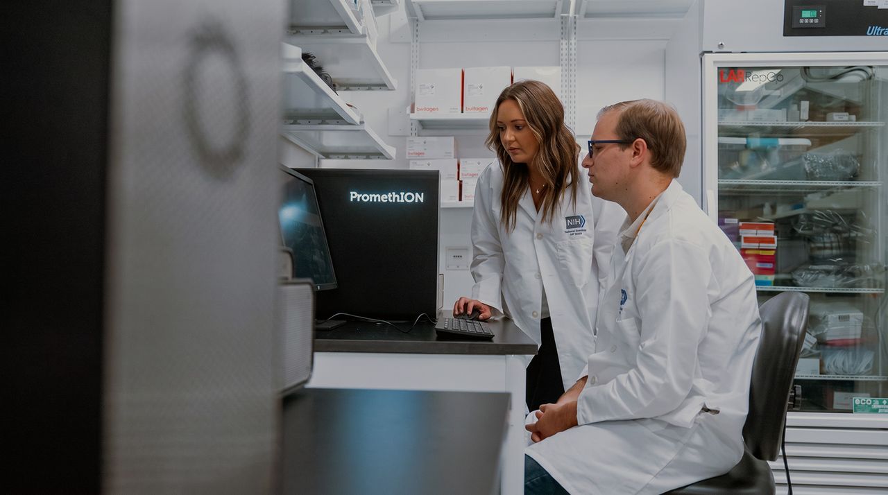 Two scientists in lab coats are working at a computer with a PromethION in a busy laboratory