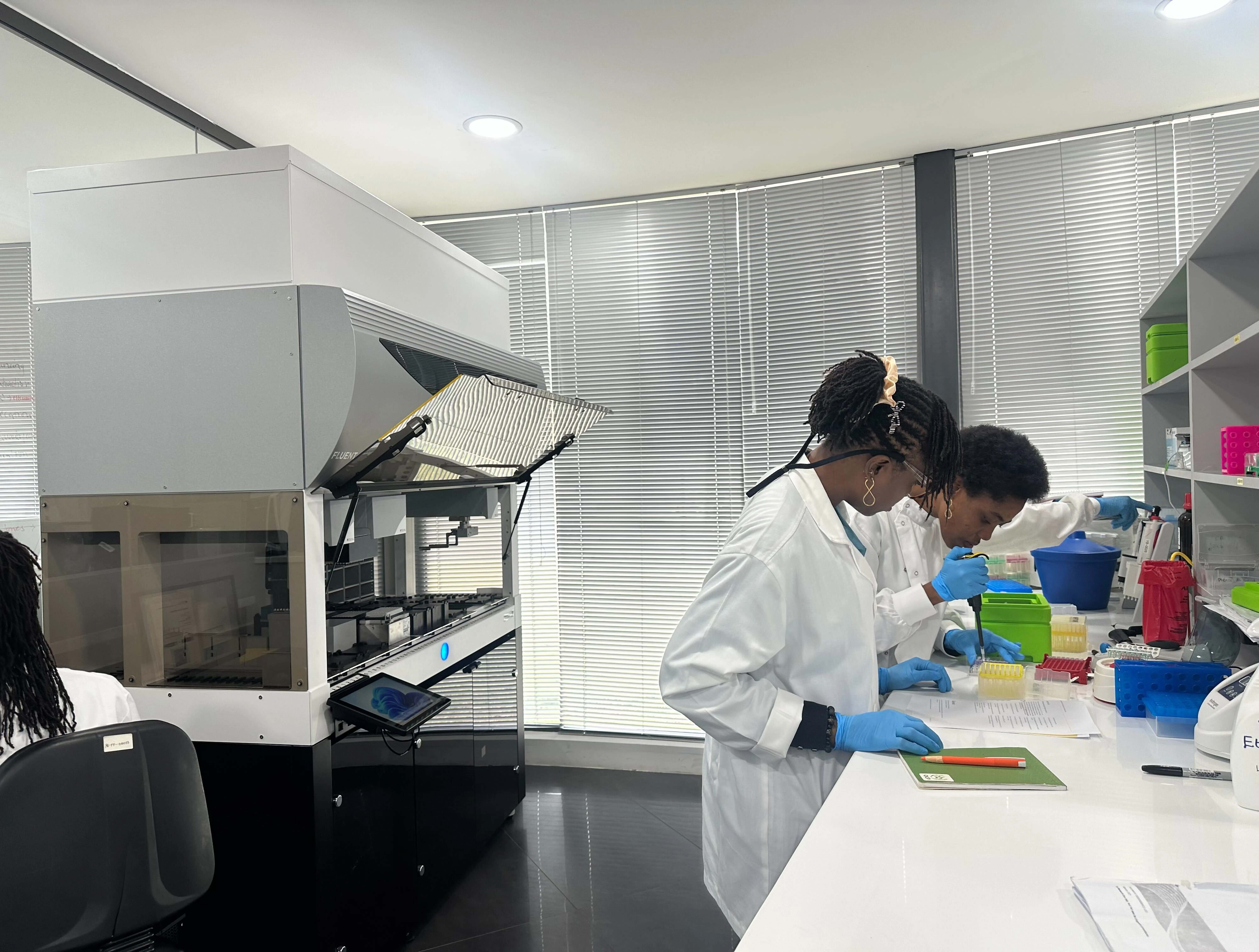Two women are stood in a lab, one of them is pipetting and the other is watching with a notebook under her hand.
