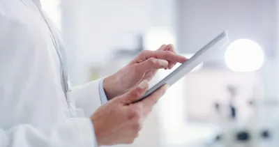 Close-up of a person in a white coat using a tablet in a bright, clinical setting.