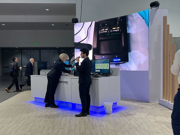 Two people stand at a conference booth, the man on the right is talking into a microphone, the women on the left is pipetting into a sequencing device